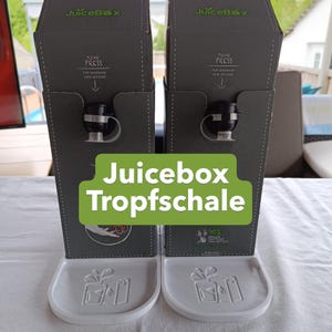 Könnte beinhalten: Zwei graue JuiceBox-Spender mit schwarzen Ausläufen und weißen Tropfschalen. Auf der Vorderseite jeder Box steht der Text JuiceBox, Please Press For Maximum One Second sowie eine Grafik mit 90% smaller footprint than bottles.