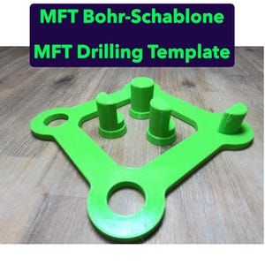 Peut inclure: Un gabarit de perçage MFT vert vif avec une forme unique, comprenant plusieurs poteaux cylindriques et une grande ouverture circulaire. Le texte "MFT Bohr-Schablone" et "MFT Drilling Template" est affiché en vert sur fond bleu foncé.