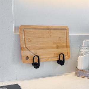 Puede incluir: Una tabla de cortar rectangular de bambú con un canal para jugos y un orificio circular en la esquina, montada en una pared mediante dos ganchos ovalados negros. La tabla presenta un acabado de madera clara y está colocada horizontalmente.