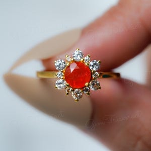 May include: A gold ring featuring a vibrant orange gemstone at its center, surrounded by a halo of sparkling white gemstones. The ring's design is reminiscent of a flower, with the gemstones arranged in a circular pattern. The ring is a piece of jewelry.