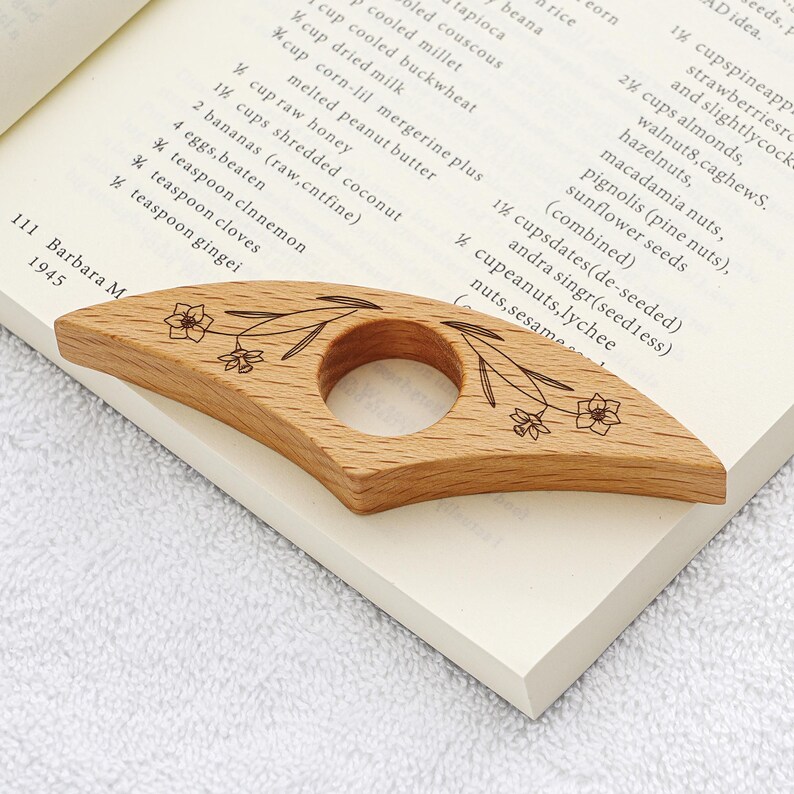 Puede incluir: Un sujetap&aacute;ginas de madera clara con un dise&ntilde;o floral descansa sobre un libro abierto. El sujetap&aacute;ginas tiene una abertura circular en el centro. Las p&aacute;ginas del libro son blancas con texto negro.