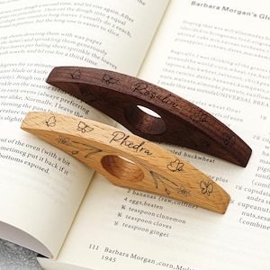 May include: Two wooden book page holders, one light and one dark, with floral and butterfly engravings. The dark holder is inscribed with "Rosalia", and the light one with "Phedra". Both have a finger hole for easy use.