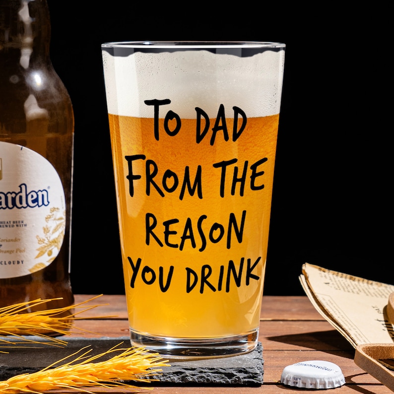 May include: A clear glass beer mug with a light yellow beer inside. The mug has black text that reads "To Dad From The Reason You Drink".
