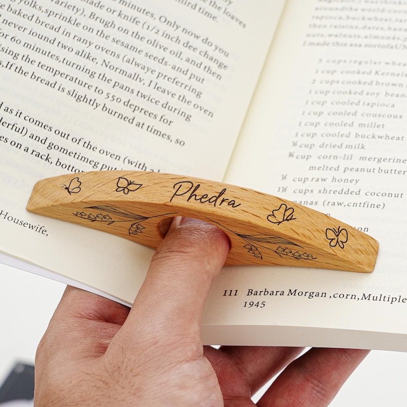 Puede incluir: Soporte de p&aacute;gina de libro de madera con el nombre "Phedra" grabado. El soporte marr&oacute;n claro presenta dise&ntilde;os de mariposas y flores. El soporte est&aacute; dise&ntilde;ado para sostenerse con un dedo.