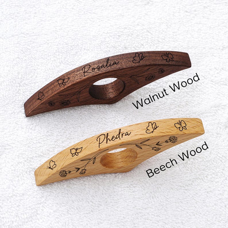 Puede incluir: Dos sujetap&aacute;ginas de madera, uno de nogal y otro de haya, con dise&ntilde;os grabados de flores y mariposas. El soporte de nogal est&aacute; inscrito con "Rosalia", y el de haya con "Phedra". Tambi&eacute;n se ve el texto "Walnut Wood" y "Beech Wood".