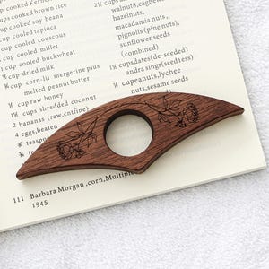 Puede incluir: Un sujetap&aacute;ginas de madera marr&oacute;n oscuro con un dise&ntilde;o floral. El marcap&aacute;ginas curvado tiene una abertura circular y detalles grabados de flores y hojas. Descansa sobre un libro abierto con texto.