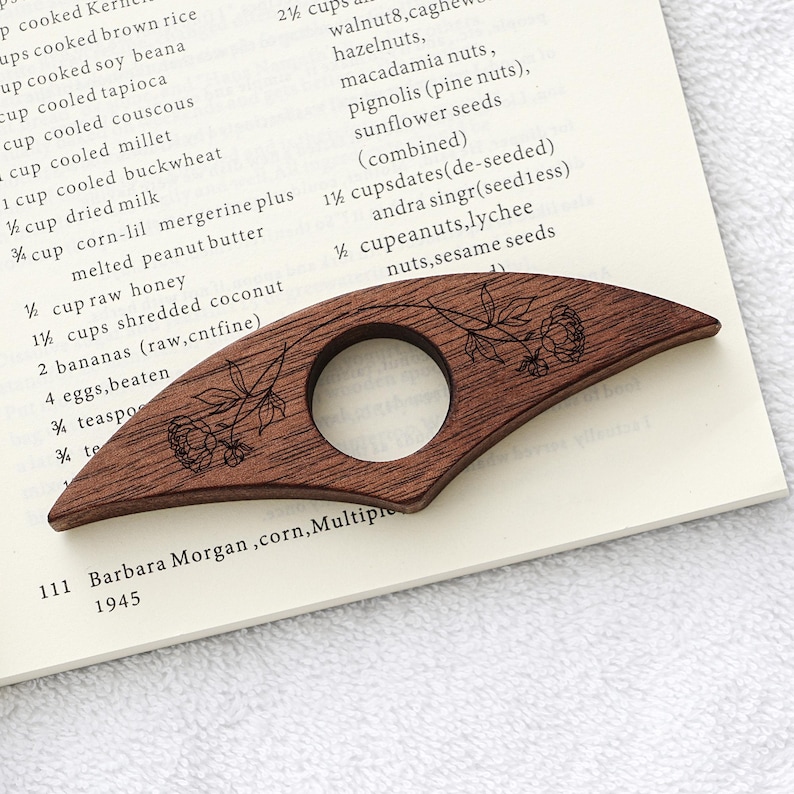 Puede incluir: Un sujetap&aacute;ginas de madera marr&oacute;n oscuro con un dise&ntilde;o floral. El marcap&aacute;ginas curvado tiene una abertura circular y detalles grabados de flores y hojas. Descansa sobre un libro abierto con texto.