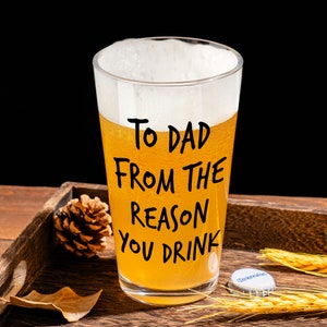 May include: A tall glass beer mug with a white head and a yellow beer inside. The mug has black writing that says "To Dad From The Reason You Drink".