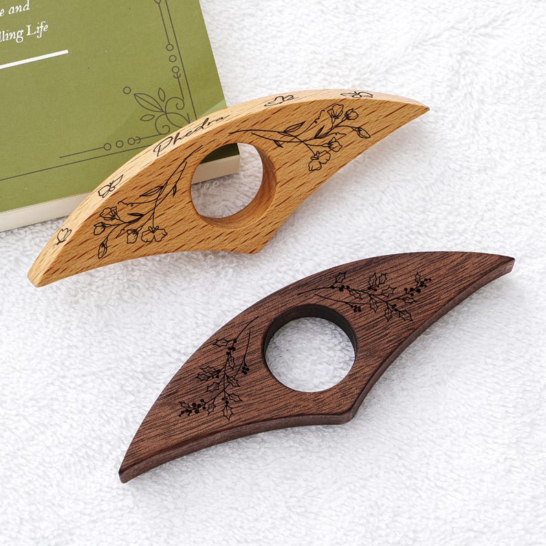 Puede incluir: Dos sujetap&aacute;ginas de madera para el pulgar. Uno es de color claro con el nombre "Phedra" y grabado floral. El otro es de color marr&oacute;n oscuro con un dise&ntilde;o floral similar. Ambos tienen un agujero circular para el pulgar y est&aacute;n dise&ntilde;ados para sujetar un libro abierto.