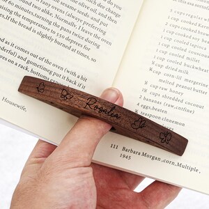 Puede incluir: Un marcap&aacute;ginas de madera marr&oacute;n oscuro con el nombre "Rosalia" grabado y dise&ntilde;os de mariposas. El marcap&aacute;ginas est&aacute; colocado en las p&aacute;ginas de un libro. El texto "111 Barbara Morgan, corn, Multiple, 1945" es visible.