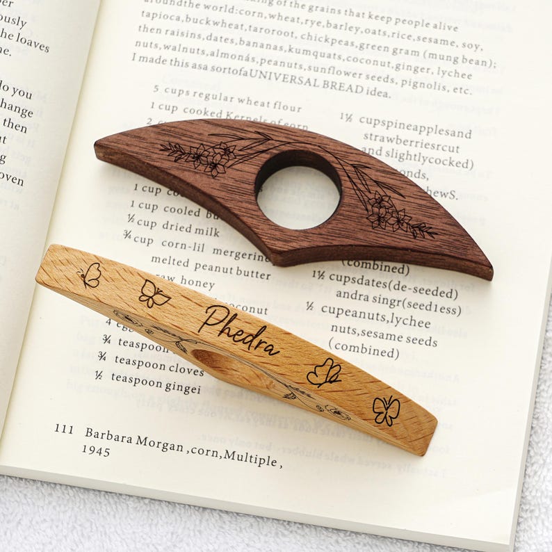 Puede incluir: Dos sujetap&aacute;ginas de madera. Uno es marr&oacute;n oscuro con grabados florales, el otro es marr&oacute;n claro con grabados de mariposas y el nombre "Phedra". Ambos tienen una abertura circular para el pulgar.