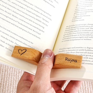 Puede incluir: Se muestran dos marcapáginas de madera, uno con un símbolo de corazón y el otro con el nombre "Rayna" grabado. Los marcapáginas son de color marrón claro y sostienen un libro abierto. Las páginas del libro son blancas con texto negro.
