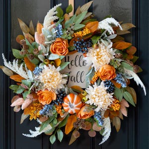 May include: A decorative autumn wreath featuring orange roses, cream chrysanthemums, blue berries, pinecones, and pampas grass. A central white sign displays the text "Hello Fall" in black script, surrounded by green and brown foliage.