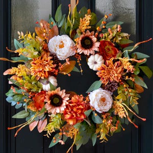May include: A circular autumn wreath featuring white peonies, orange dahlias, sunflowers, and small white pumpkins. The arrangement includes green foliage, brown pinecones, and orange berry sprigs, creating a dense, multi-textured floral display.