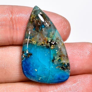 Puede incluir: Una gema pulida con forma de lágrima, con una base azul turquesa vibrante que se mezcla con una combinación de gris, marrón y negro en la parte superior. La piedra tiene un acabado brillante, resaltando sus patrones naturales.