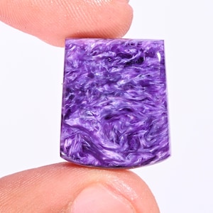 Op de afbeelding: Een gepolijste, rechthoekige edelsteen met een afgeronde onderrand. De steen vertoont een wervelend patroon van diep paars en wit, waardoor een marmerachtig effect ontstaat. De edelsteen wordt tussen twee vingers tegen een witte achtergrond gehouden.