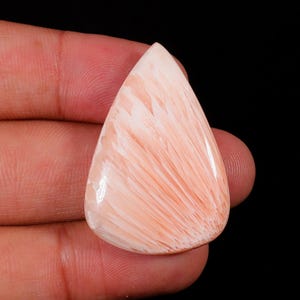 Può includere: Una pietra preziosa lucidata a forma di pera con una delicata tavolozza di colori pesca e bianco. La pietra presenta un distinto motivo fibroso radiante che crea un effetto setoso e chatoyant sulla sua superficie liscia e arrotondata.