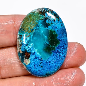 Puede incluir: Una gema ovalada con una llamativa mezcla de colores. La piedra presenta una base turquesa vibrante, acentuada con parches de azul profundo, verde y marrón. La superficie pulida refleja la luz, resaltando los patrones y texturas naturales.