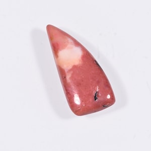 May include: A polished, elongated gemstone with a rounded tip. The stone features a blend of pink, white, and peach hues, with darker pink areas and small black inclusions. The gemstone is smooth and glossy.