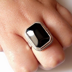 Könnte beinhalten: Ein silberner Ring mit einem schwarzen rechteckigen Edelstein, der in einem dekorativen Rand gefasst ist.