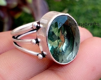 Könnte beinhalten: Ein Silberring mit einem großen, ovalen grünen Edelstein. Der Ring hat ein geteiltes Schaftdesign mit dekorativen Akzenten. Der Edelstein ist in einer Lünettenfassung gefasst. Der Ring wird vor einem natürlichen Hintergrund präsentiert.