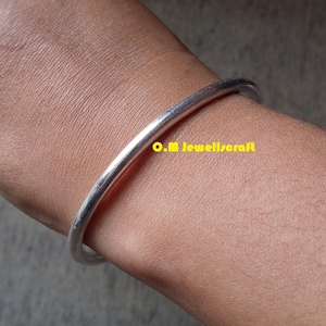 Puede incluir: Un brazalete de color plateado. El brazalete redondo y pulido se lleva en el brazo de una persona. El texto "O.M jewell/craft" es visible en amarillo. El brazalete es una joya sencilla y elegante.
