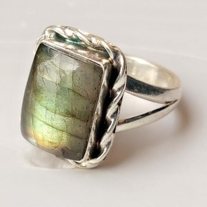 May include: A silver ring featuring a rectangular, iridescent labradorite gemstone. The stone displays shades of green, yellow, and blue. The ring has a braided design around the stone and a split shank.