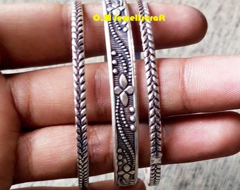 May include: Three silver-toned metal bangles featuring oxidized detailing. The two outer bangles have a repeating chevron pattern, while the wider center bangle displays a floral and dotted motif. The text O.M Jewellcraft is visible.