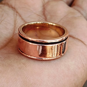 May include: A copper-colored ring with a black band and etched details. The ring is a wide band style, with a polished finish. The ring is shown on a hand.
