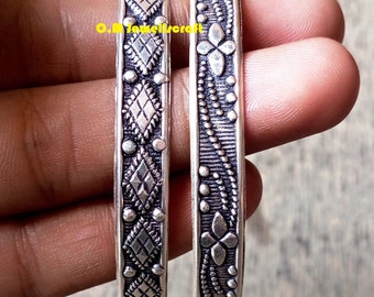 May include: Two silver bangles with intricate designs. One bangle features a repeating diamond pattern, while the other has a floral motif.