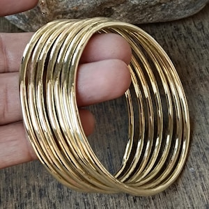 Pode incluir: Um conjunto de dez pulseiras de bracelete douradas. As pulseiras estão empilhadas umas sobre as outras e todas são do mesmo tamanho.