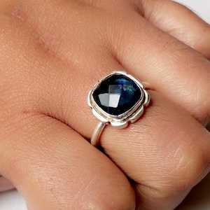 Könnte beinhalten: Ein silberner Ring mit einem quadratischen, facettierten, dunkelblauen Edelstein. Der Edelstein ist in einer dekorativen, geschwungenen Lünette gefasst. Der Ring wird an einer Hand gezeigt, um das Design und die Farbe des Schmucks hervorzuheben.