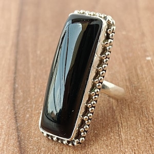 May include: A silver ring featuring a large, rectangular black onyx stone. The stone is polished and reflective, set within a silver frame with a beaded border. The ring is displayed on a wooden surface.