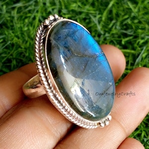May include: A silver ring with a large oval-shaped blue labradorite gemstone. The ring has a detailed silver band with a braided design.