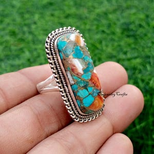 Puede incluir: Un anillo de plata con una piedra grande, rectangular, de turquesa y cobre. La piedra tiene una mezcla de colores azul, naranja y blanco. El anillo tiene un borde plateado detallado y texturizado. El anillo se sostiene en una mano sobre un fondo verde.