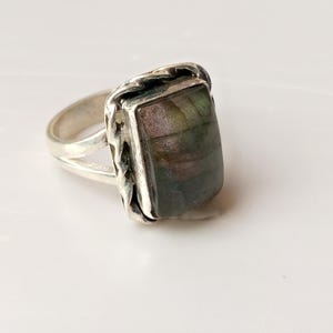 Op de afbeelding: Een zilveren ring met een rechthoekige, iriserende edelsteen. De steen toont tinten groen, paars en bruin. De ring heeft een gespleten schacht en een getextureerde bezel setting, wat een vintage of ambachtelijke uitstraling cre&euml;ert.