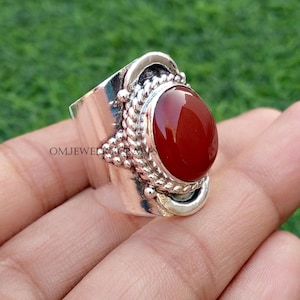 May include: A silver ring with a large, oval, red gemstone. The ring has a wide band with decorative silver accents, including a braided rope design and small silver beads. The ring is shown on a hand against a blurred green background.