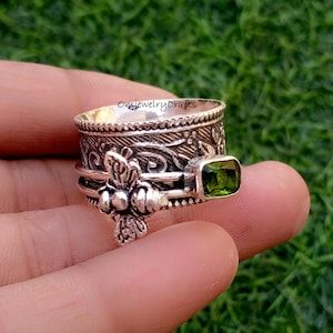 Può includere: Un anello color argento con un design ornato e una gemma verde. L'anello ha una fascia rotante e un dettaglio floreale. Il testo "omjewelrycrafts" è visibile sull'anello.