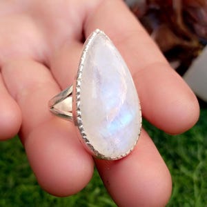 Puede incluir: Anillo de plata con una piedra lunar en forma de pera. La piedra iridiscente muestra un color blanco nacarado con destellos azules y morados. El engaste del anillo tiene un borde de cuentas.