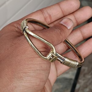 K&ouml;nnte beinhalten: Ein goldfarbenes Armreif-Armband mit einem einzigartigen, gedrehten Drahtdesign und einem Verschluss, der einem Haken &auml;hnelt.