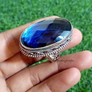 Op de afbeelding: Een opvallende ring met een grote, ovaal geslepen, diepblauwe edelsteen. De ring heeft een zilverkleurige metalen zetting met gedetailleerd filigraanwerk rond de steen en op de band. De ring wordt in een hand gehouden tegen een groene achtergrond.