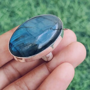 May include: A silver ring featuring a large, oval-shaped labradorite stone. The stone displays a shimmering blue and teal color, set in a polished silver band. The ring is shown on a hand against a blurred green background. The text "Omjewelry Crafts" is visible.
