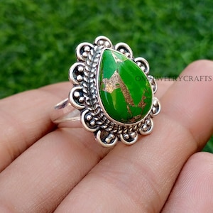 Puede incluir: Anillo de plata con una piedra preciosa verde brillante en forma de pera con inclusiones doradas. La piedra está engastada en un bisel decorativo y festoneado. El anillo se muestra sobre un fondo verde. Joya.