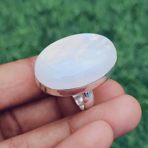 May include: A large, oval-shaped moonstone ring with a polished, iridescent surface. The gemstone is set in a silver band. The ring is displayed against a blurred green background.