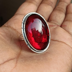 May include: A silver ring featuring a large, oval-shaped, red gemstone set in a bezel setting. The ring has a decorative border around the gemstone.