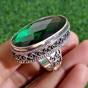 May include: A silver ring with an oval, faceted, emerald-green gemstone. The ring has an ornate, scrollwork design around the gemstone and on the band. The text "Jewelry Crafts" is visible on the ring.