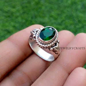 Könnte beinhalten: Ein silberner Ring mit einem ovalen, smaragdgrünen Edelstein. Der Ring hat ein geflochtenes Seildesign um den Edelstein und dekorative Akzente an den Seiten. Der Text "OMJEWELRYCRAFTS" ist auf der Seite sichtbar.