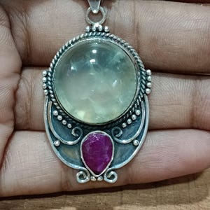 May include: A silver pendant with a large, round, clear green stone and a smaller, teardrop-shaped, pink stone.