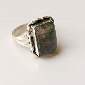 Op de afbeelding: Een zilveren ring met een rechthoekige, iriserende edelsteen. De steen toont tinten groen, paars en bruin. De ring heeft een gespleten schacht en een getextureerde bezel setting, wat een vintage of ambachtelijke uitstraling cre&euml;ert.
