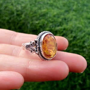 May include: A silver ring with an oval, amber-colored gemstone. The gemstone is set in a decorative bezel with a twisted rope design. The ring's band features ornate scrollwork on the sides, creating a vintage aesthetic.